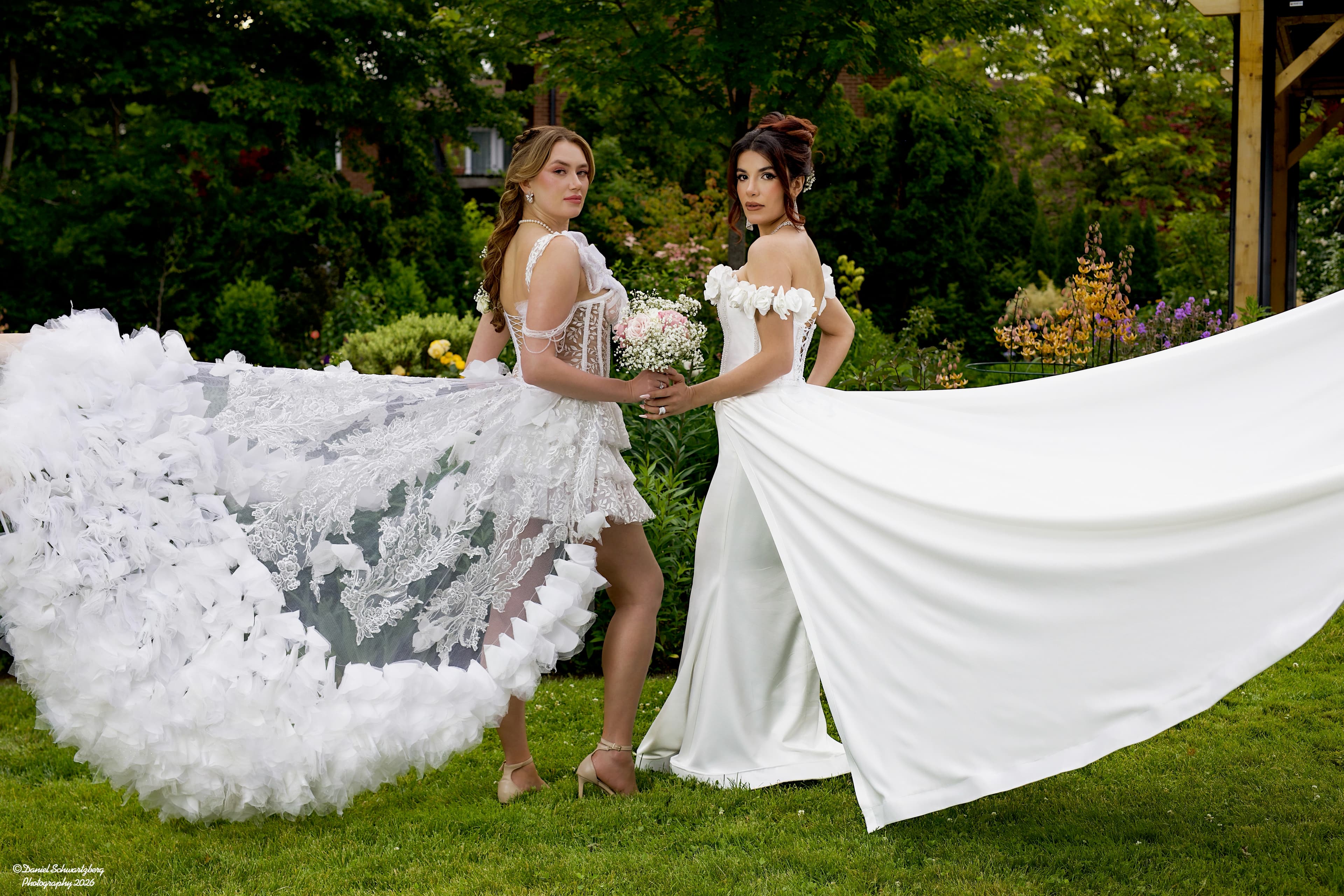 Two brides in couture white gowns with sweeping trains in a lush garden, one holding a pastel rose bouquet